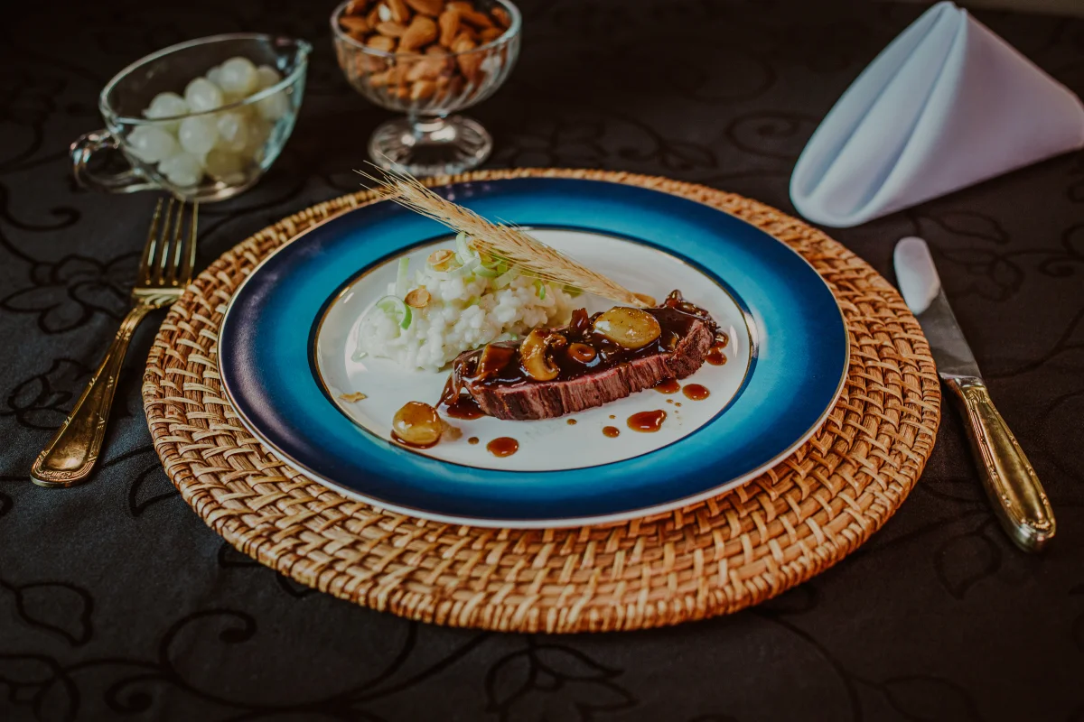 Prato gourmet com bife ao molho, risoto branco e decoração com espiga de trigo em prato azul com borda dourada