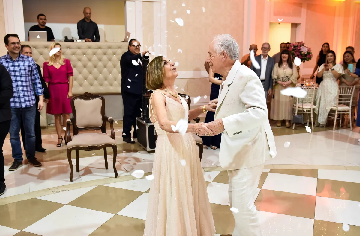 Casal idoso dançando em festa elegante com convidados ao fundo e pétalas caindo