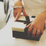 Mãos femininas segurando caixa de presente com laço marrom sobre vestido branco com detalhes em renda
