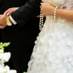Noiva segurando terço de pérolas com vestido branco detalhado e noivo ao lado com terno preto e relógio dourado
