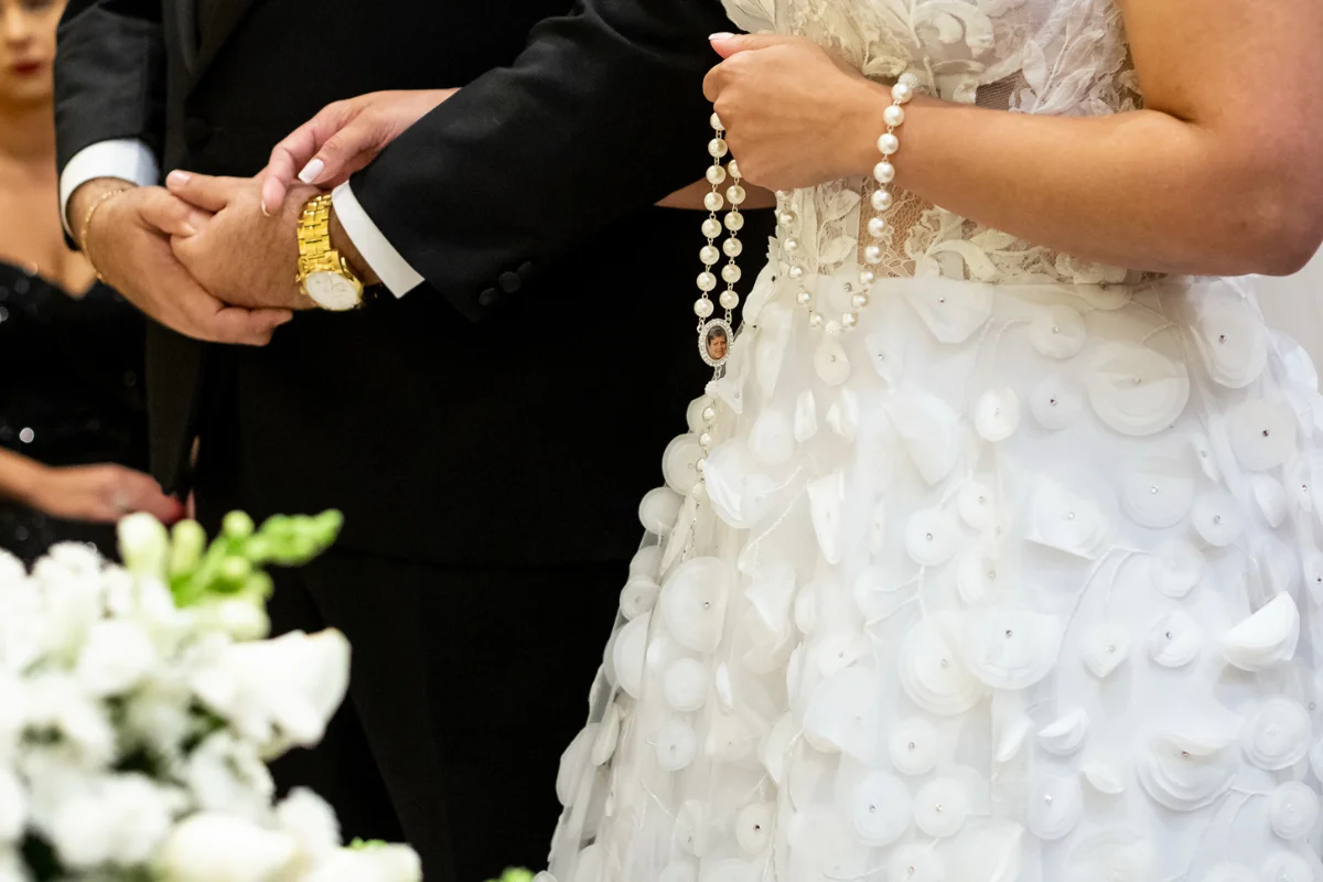 Noiva segurando terço de pérolas com vestido branco detalhado e noivo ao lado com terno preto e relógio dourado