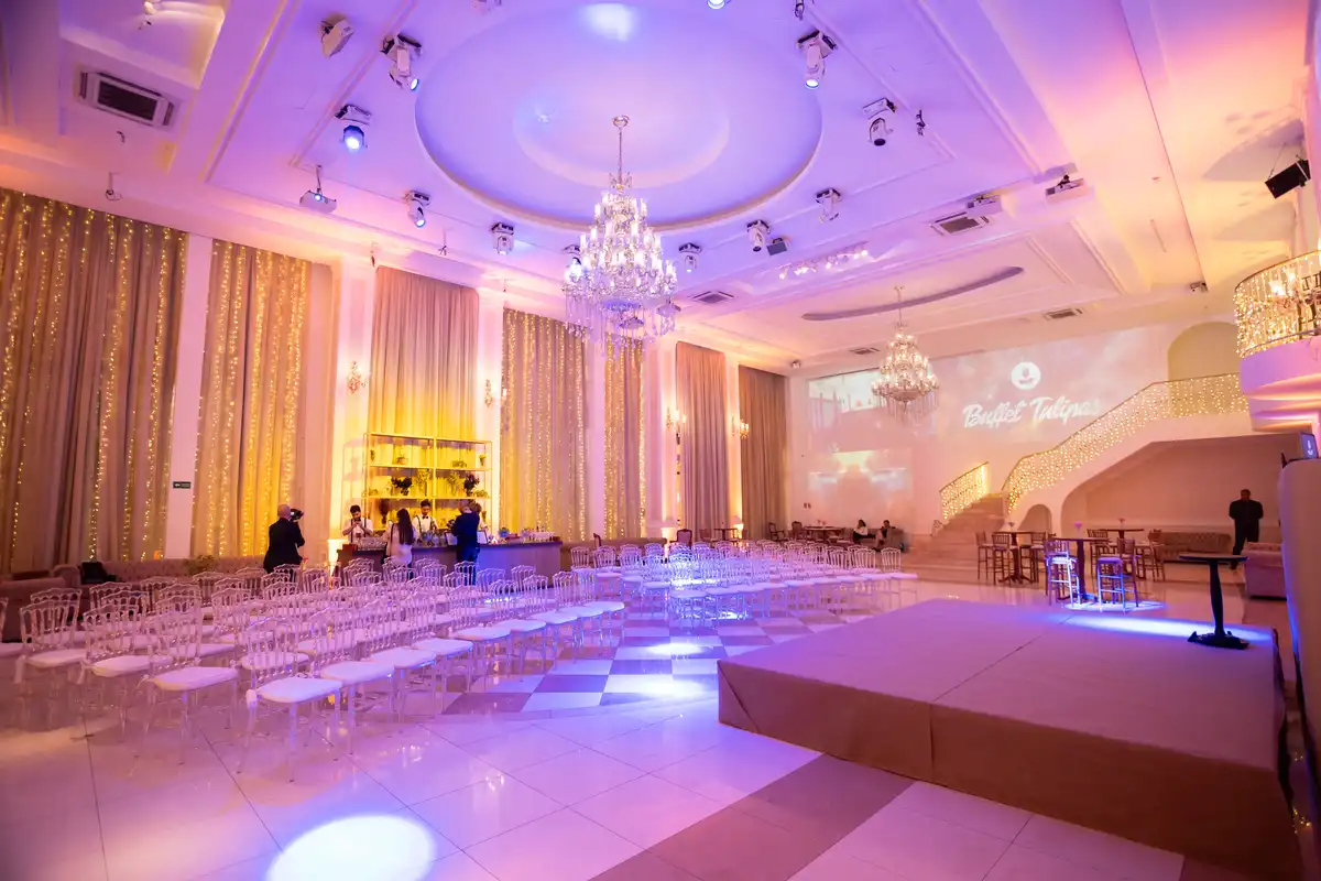 Salão de festa elegante com iluminação suave, cortinas douradas, lustres de cristal e uma área de palco preparada para eventos.