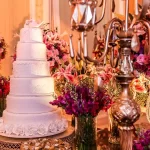 Como escolher a decoração de casamento que mais combina com você? Bolo de casamento branco de cinco andares decorado com flores brancas e topo de pombas, cercado por arranjos florais em tons de rosa, lilás e roxo em um ambiente luxuoso