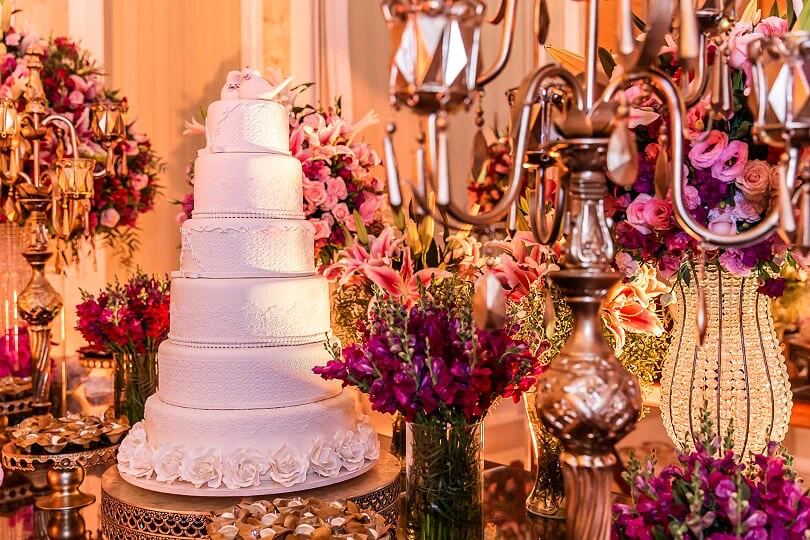 Bolo de casamento branco de cinco andares decorado com flores brancas e topo de pombas, cercado por arranjos florais em tons de rosa, lilás e roxo em um ambiente luxuoso
