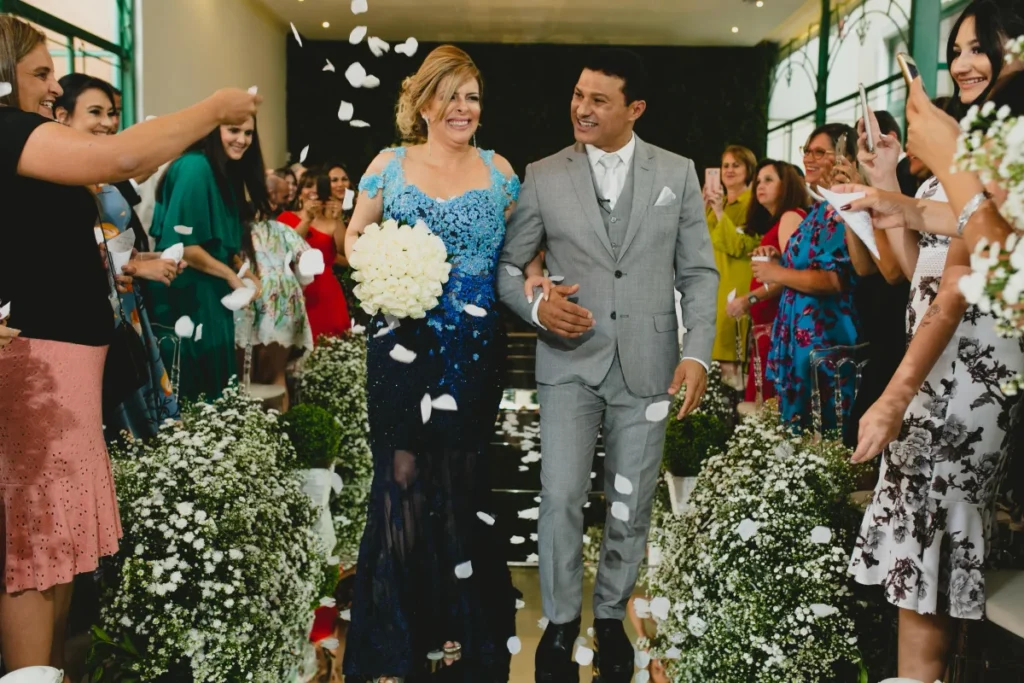 Casal feliz caminhando pelo corredor decorado com flores brancas e convidados jogando pétalas em cerimônia de casamento