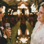 Votos de casamento: como fazer e quando renovar? Casal de noivos sorrindo durante casamento ao ar livre, com iluminação de velas ao redor e reflexo na água ao fundo, celebrando amor e união.