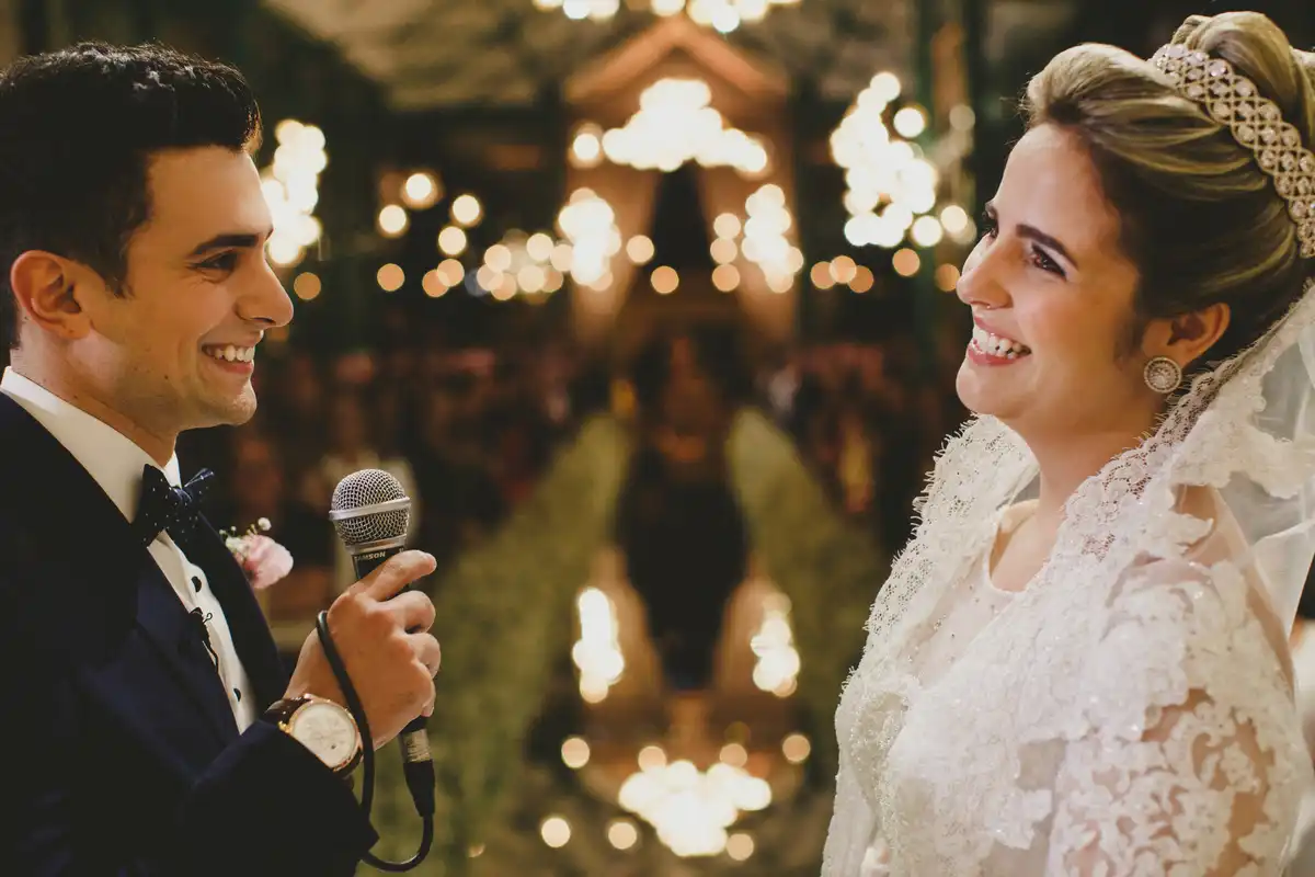 Casal de noivos sorrindo durante casamento ao ar livre, com iluminação de velas ao redor e reflexo na água ao fundo, celebrando amor e união.