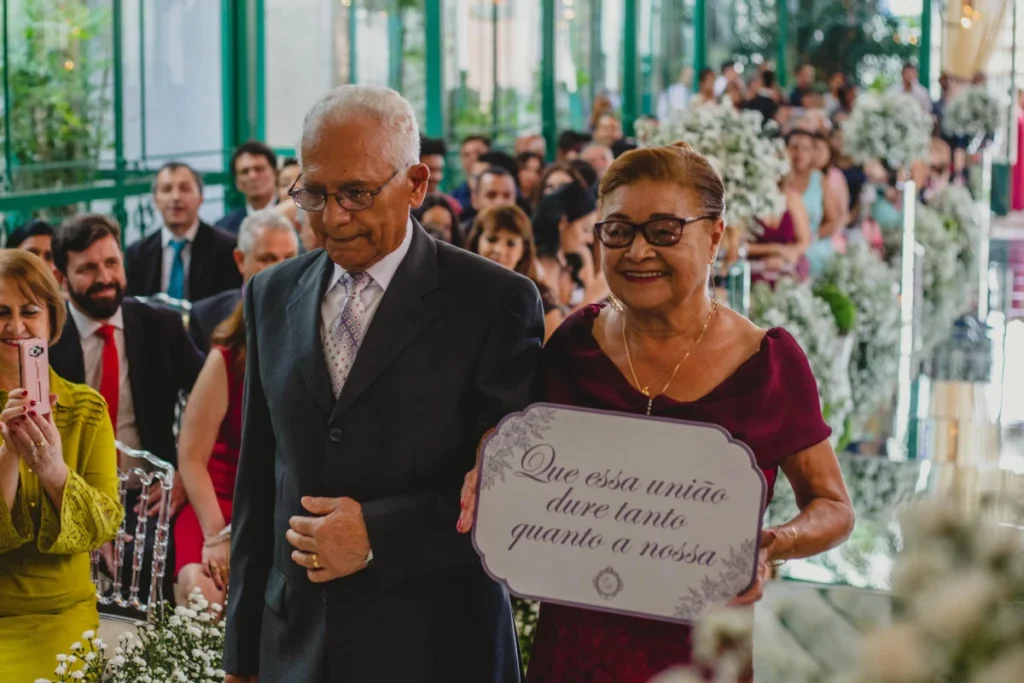 Casal idoso elegante caminhando em cerimônia de casamento com placa escrita "Que essa união dure tanto quanto a nossa"