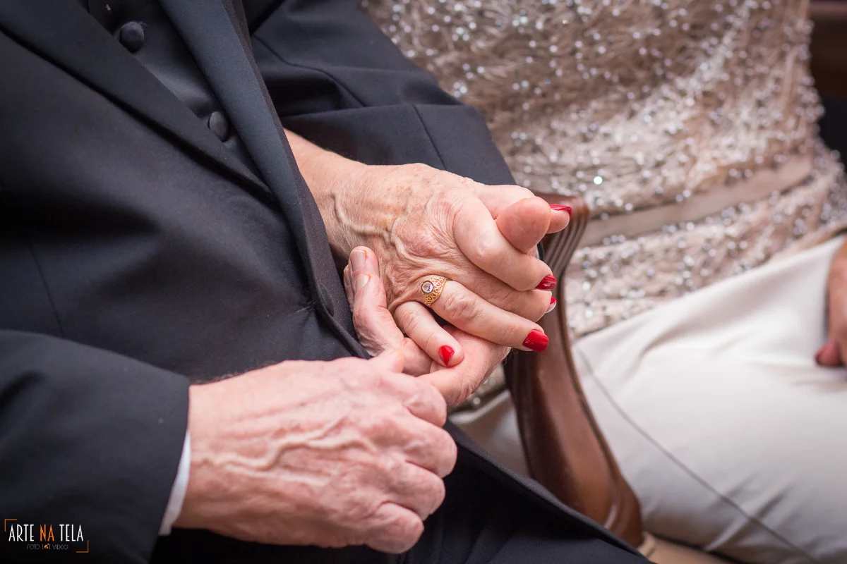 Casal idoso de mãos dadas, mostrando anel dourado e unhas vermelhas, com roupas formais