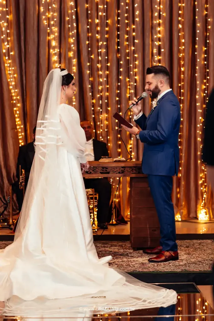 Casamento com cerimônia tradicional, noiva e noivo trocando votos em ambiente decorado com luzes douradas e cortinas de tecido, com convidados ao fundo.