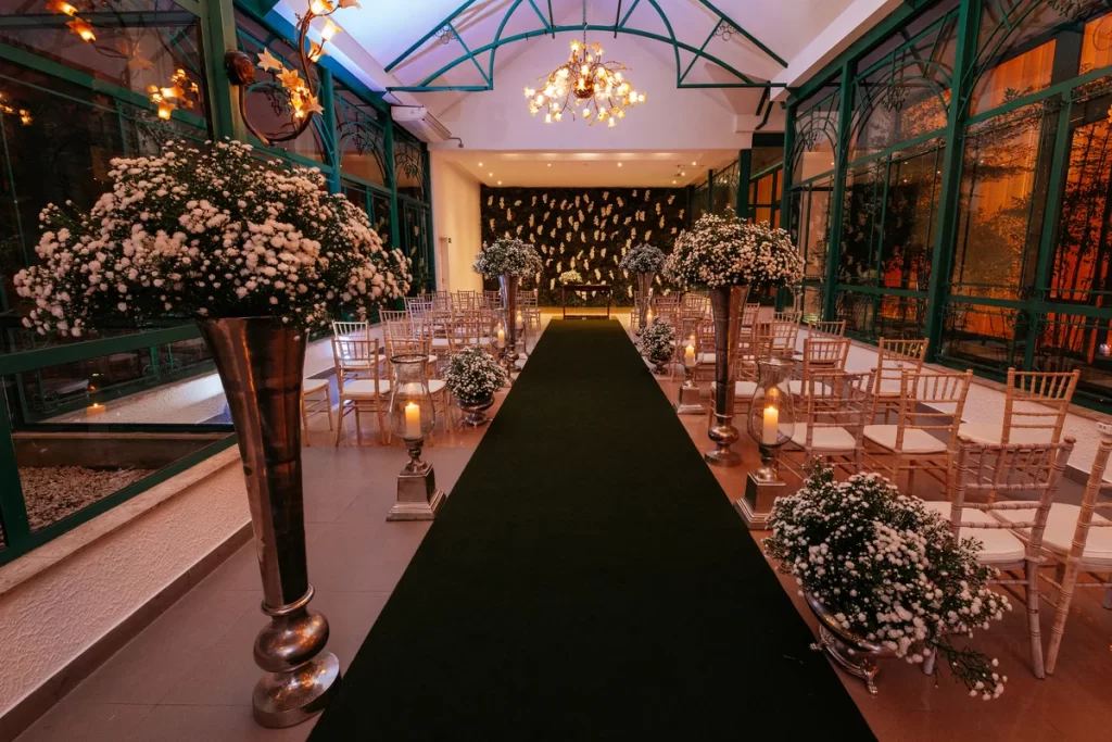 Corredor de cerimônia de casamento decorado com flores brancas e velas em castiçais, cadeiras de madeira claras alinhadas ao lado