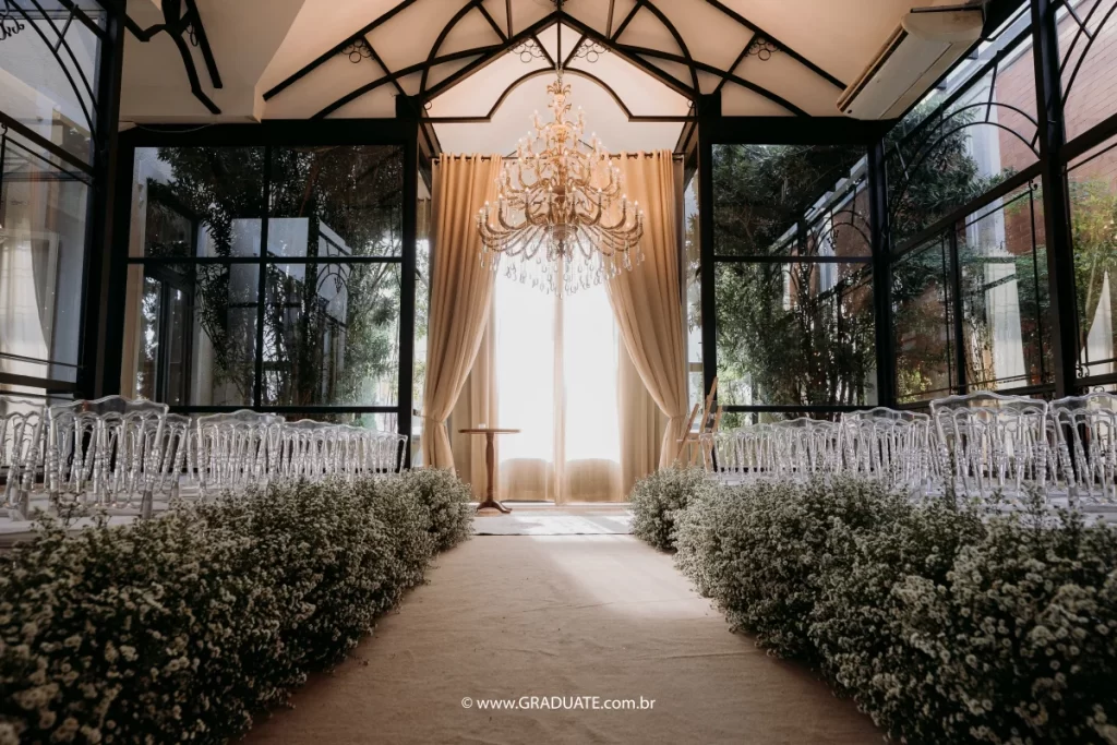 corredor decorado com flores brancas para cerimônia de casamento em salão com cadeiras transparentes e lustre de cristal