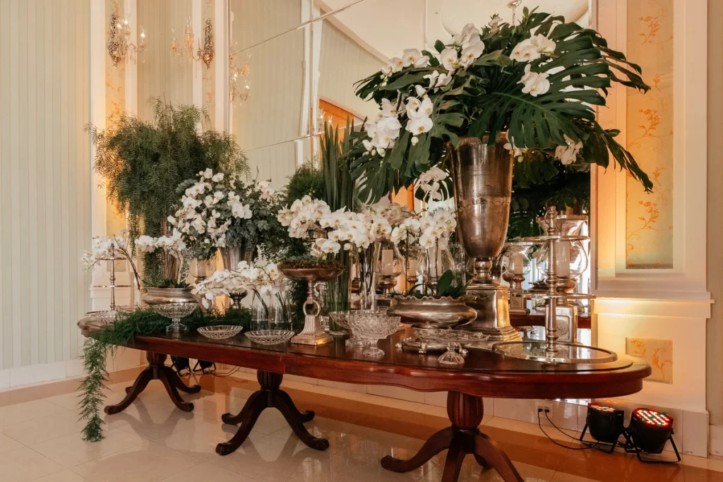 Mesa de madeira com arranjos florais brancos e folhagens verdes em vasos prateados em ambiente elegante com espelhos e iluminação quente