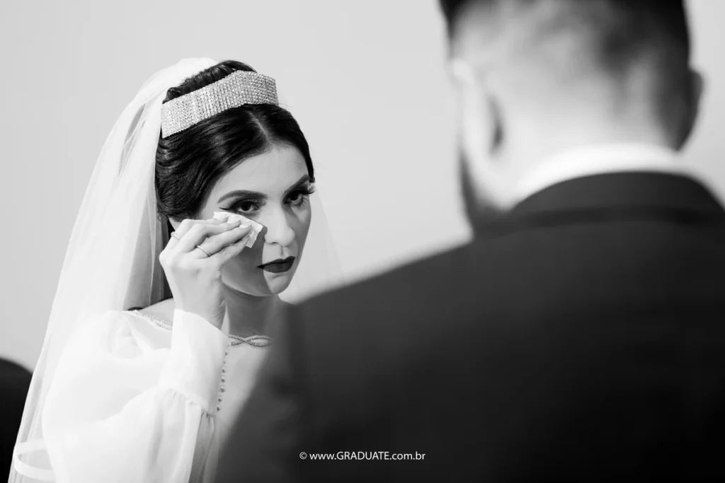Noiva enxugando lágrimas durante cerimônia de casamento civil, com expressão emocional e maquiagem marcante, vestida de noiva com véu e tiara.