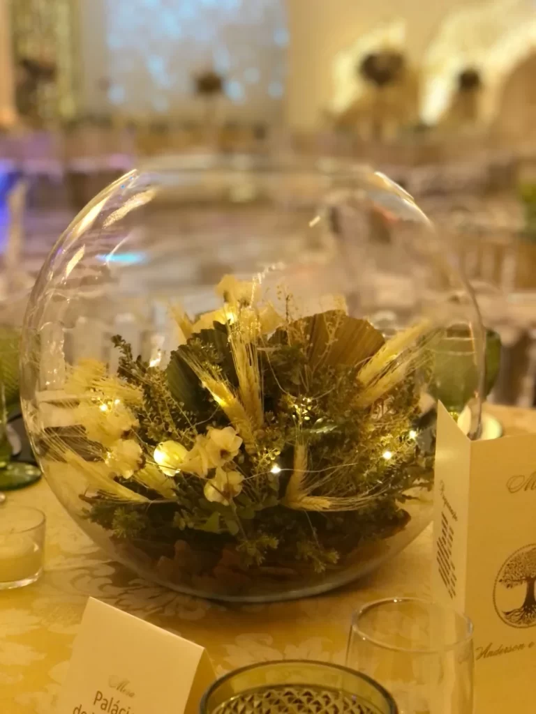 Arranjo de flores secas e luzes decorativas dentro de vaso de vidro redondo em mesa de evento