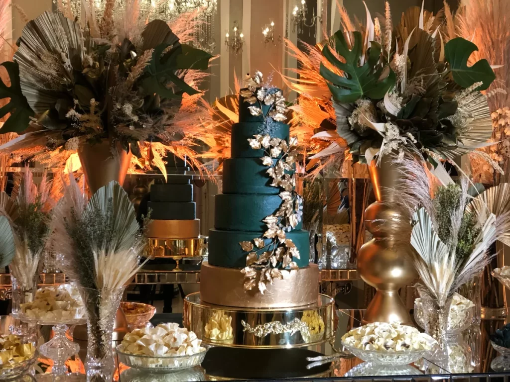 Bolo de casamento verde escuro com detalhes dourados e decoração de folhas douradas, rodeado por arranjos florais secos e vasos dourados em mesa espelhada