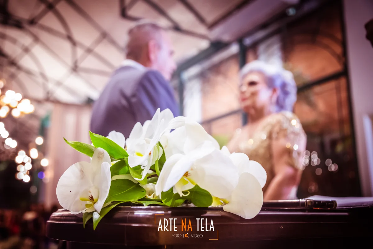 Buquê de flores brancas em foco com casal de idosos desfocado ao fundo em ambiente interno decorado