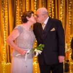 Bodas de cristal: como comemorar os 15 anos juntos Casal idoso elegante se beijando em evento com decoração de luzes amarelas e músico tocando saxofone ao fundo