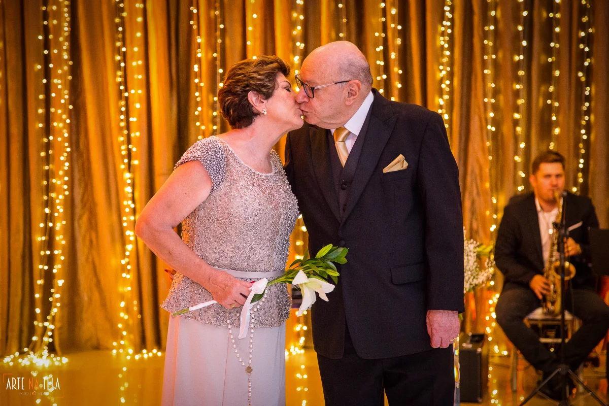 Casal idoso elegante se beijando em evento com decoração de luzes amarelas e músico tocando saxofone ao fundo
