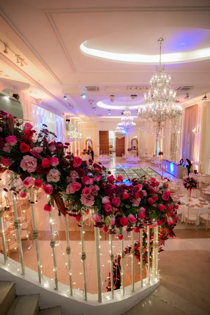 Decoração com flores rosas e vermelhas em escada iluminada em salão de festa elegante com lustres de cristal e mesas decoradas