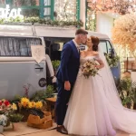 O que significa um casamento de luxo de verdade? Noivos se beijando em frente a uma van vintage decorada com flores coloridas e luzes em ambiente de casamento