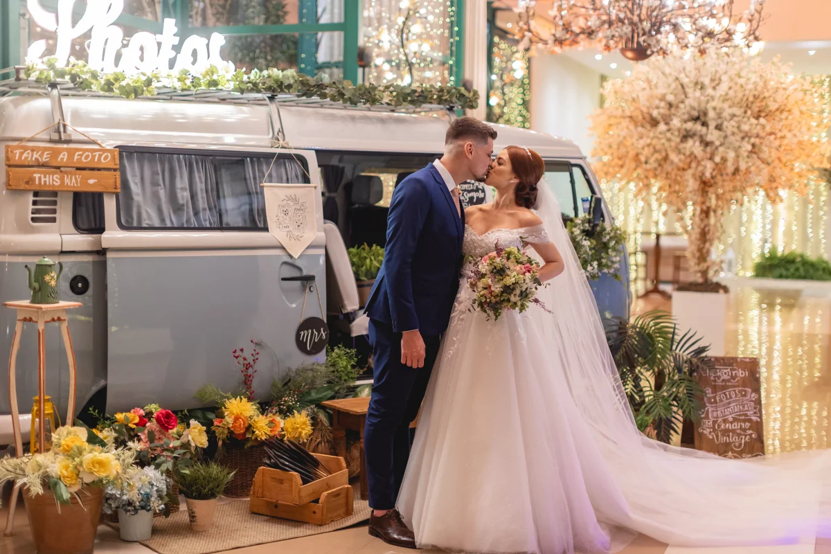Noivos se beijando em frente a uma van vintage decorada com flores coloridas e luzes em ambiente de casamento