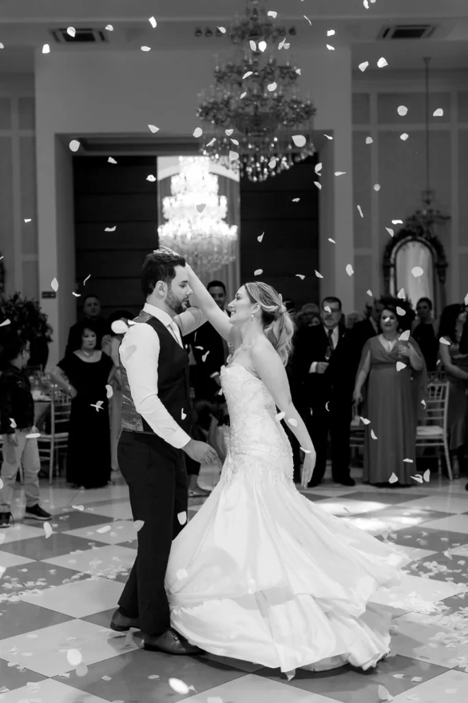 Casal de noivos dançando na pista de casamento com convidados ao fundo, confetes no ar e decoração elegante no salão.
