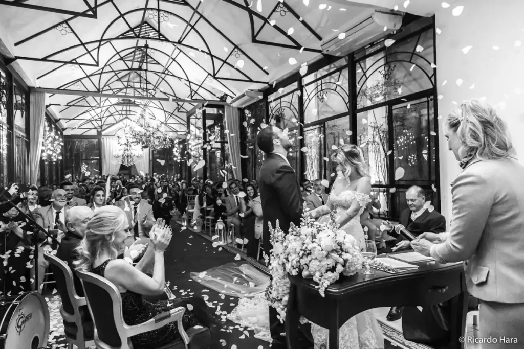 Casamento celebrado em um salão elegante com convidados e decoração sofisticada. Fotografia em preto e branco.