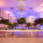 Decoração para eventos corporativos: veja 3 ideias para se inspirar Decoração elegante para eventos corporativos em salão sofisticado com iluminação colorida, cristaleira e arranjos florais decorativos.