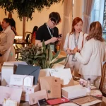 Grupo conversando em evento de negócios, com decoração elegante e diversidade de produtos em destaque na mesa.