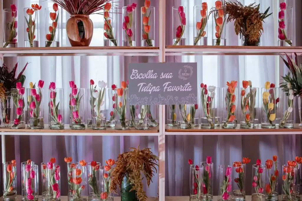 mostruario tulipas cores varidas Mostruário de taças de vidro com tulipas de cores variadas, como rosa, laranja e amarelo, em uma feira ou evento, com uma placa que diz 'Escolha sua Tulipa Favorita' ao fundo.
