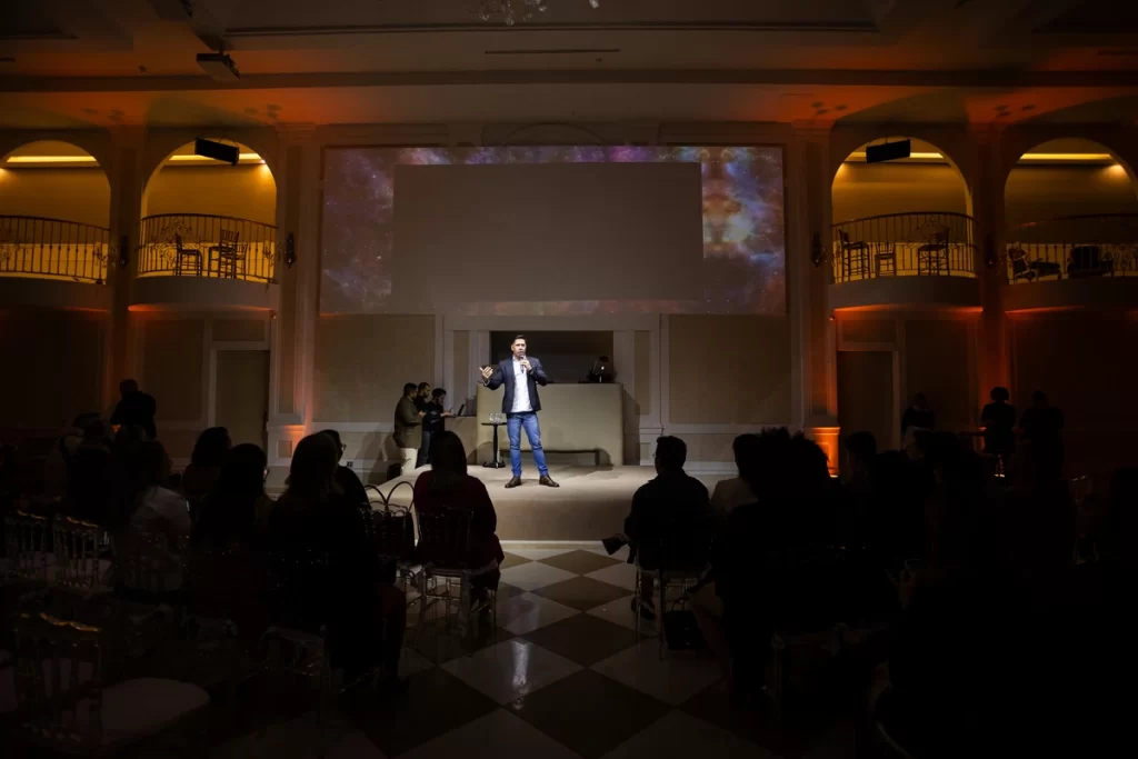 palestra conferencia evento corporativo Homem apresentando em palco iluminado, com público assistindo em um ambiente indoor decorado com iluminação quente e projeções na parede ao fundo.
