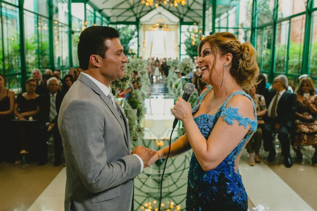Noiva com microfone na mão de frente para noivo durante cerimônia de casamento em salão decorado com flores brancas