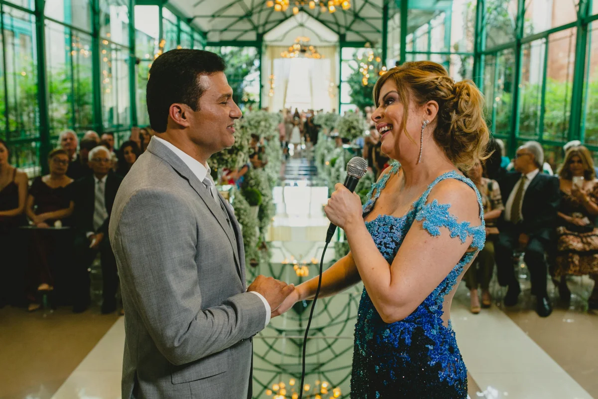 Noiva com microfone na mão de frente para noivo durante cerimônia de casamento em salão decorado com flores brancas