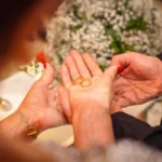 Bodas de ouro: ideias de decoração e rituais emocionantes Mãos segurando alianças de casamento douradas com fundo desfocado de flores brancas