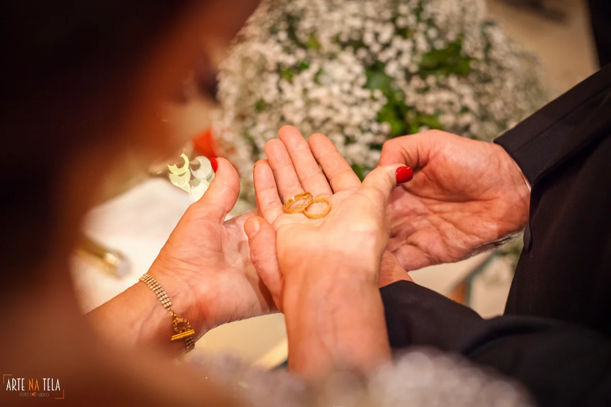 Mãos segurando alianças de casamento douradas com fundo desfocado de flores brancas
