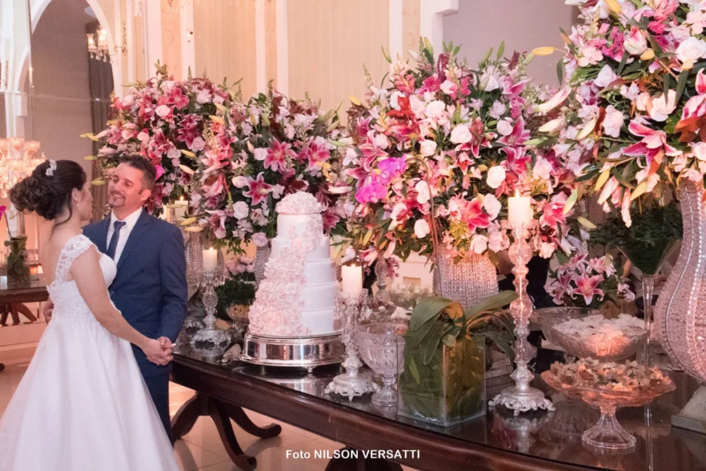 Noivos posando ao lado de mesa com bolo de casamento e arranjos florais com rosas lilases.