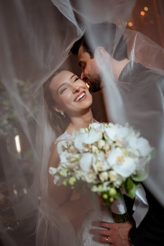 Casal de noivos sorrindo e abraçando sob o véu de casamento com buquê de flores brancas