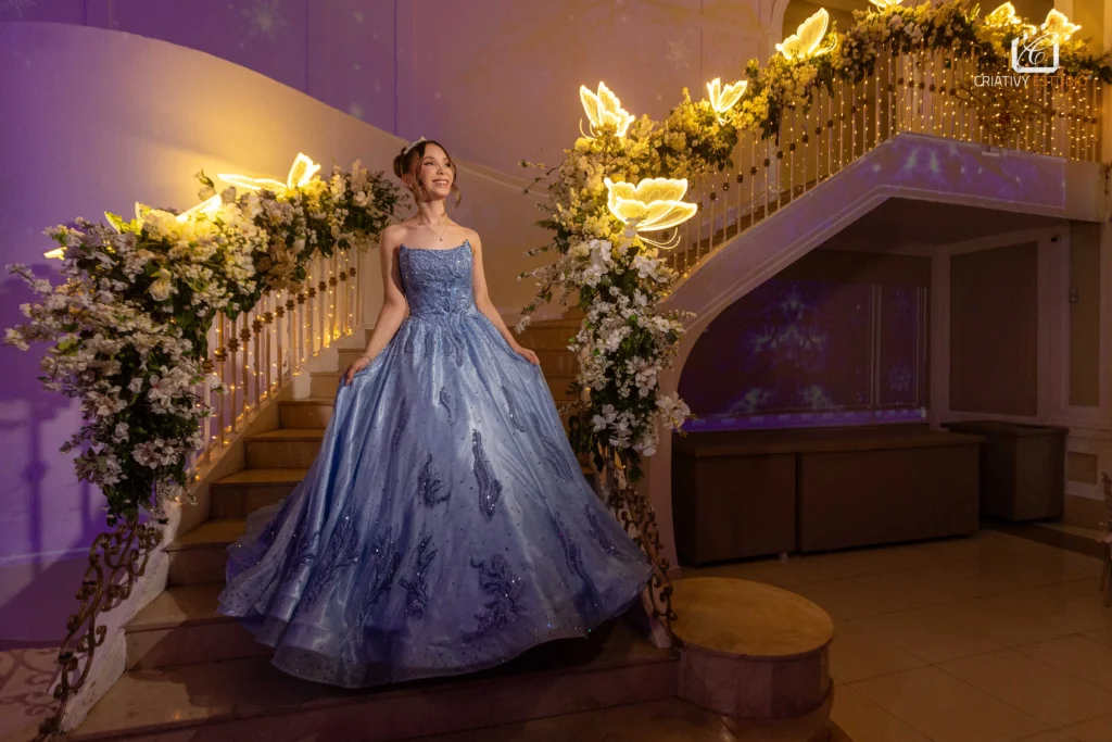Debutante com vestido azul deslumbrante em escadaria decorada com flores e luzes em festa de 15 anos