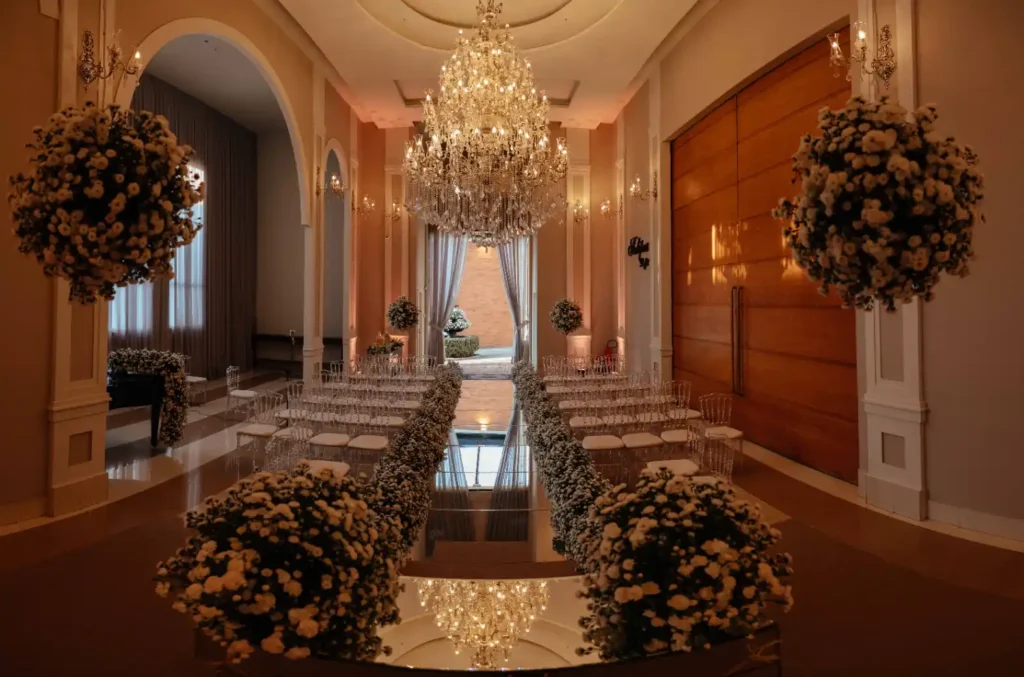 Decoração de casamento elegante em salão clássico com mesas espelhadas, arranjos de flores brancas e escuras, lustres de cristal e corredor central com iluminação dourada