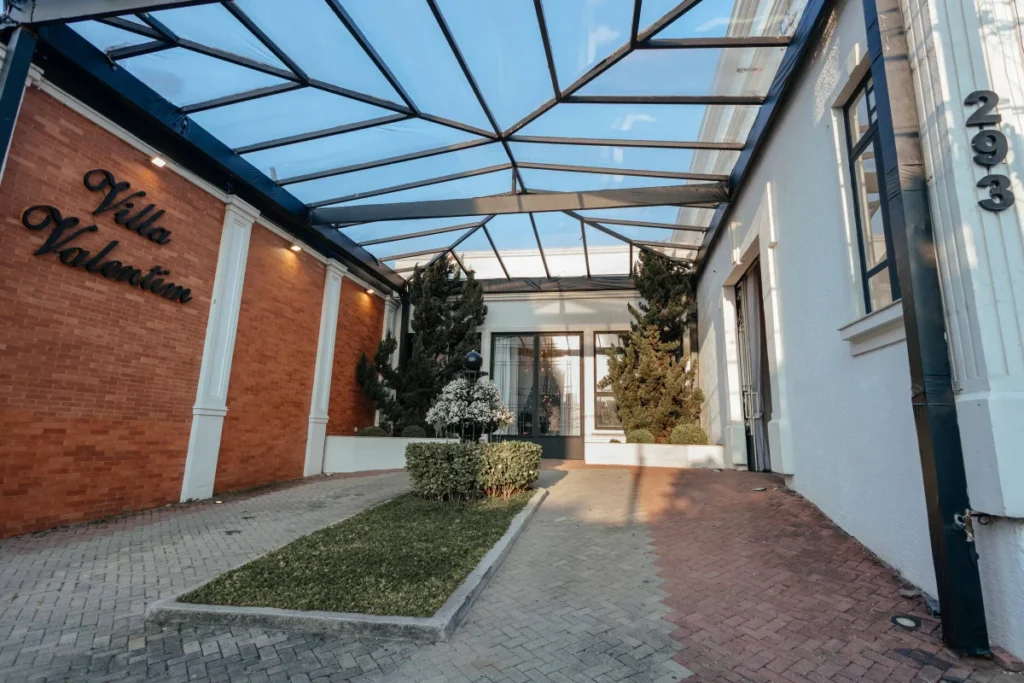 Entrada da Villa Valentin com parede de tijolos, jardim central e cobertura de vidro