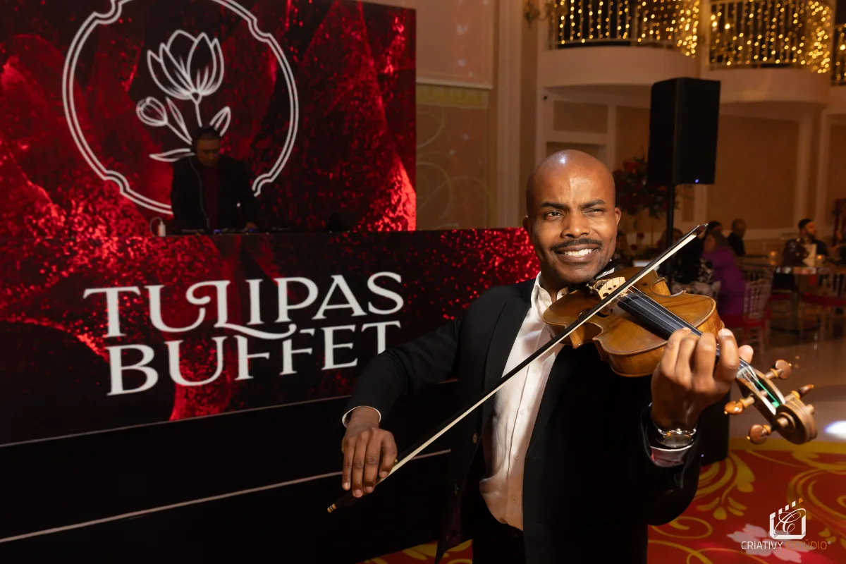 Músico tocando violino em evento elegante com decoração e painel de fundo escrito Tulipas Buffet