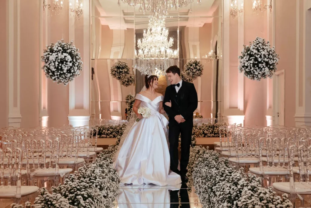 Noivos sorrindo caminhando pelo corredor decorado com flores brancas em casamento elegante