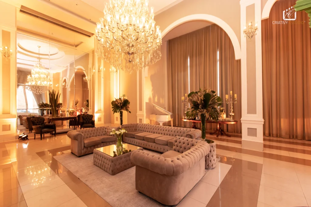Sala de estar luxuosa com sofá em formato de U, lustre de cristal, cortinas bege e decoração elegante com plantas e velas
