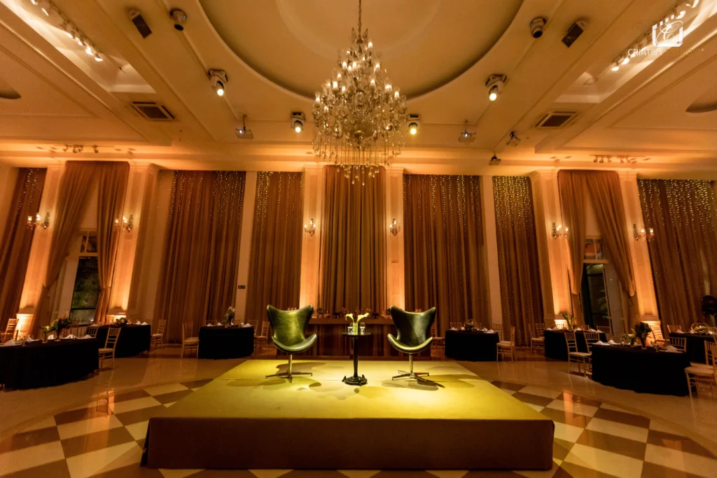 Sala de evento elegante com cortinas douradas, lustre de cristal e duas poltronas verdes em destaque