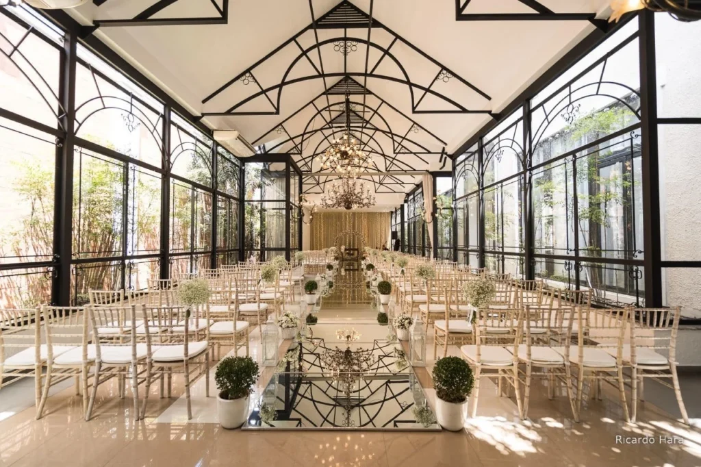 Salão de casamento decorado com cadeiras brancas, plantas em vasos e lustres elegantes