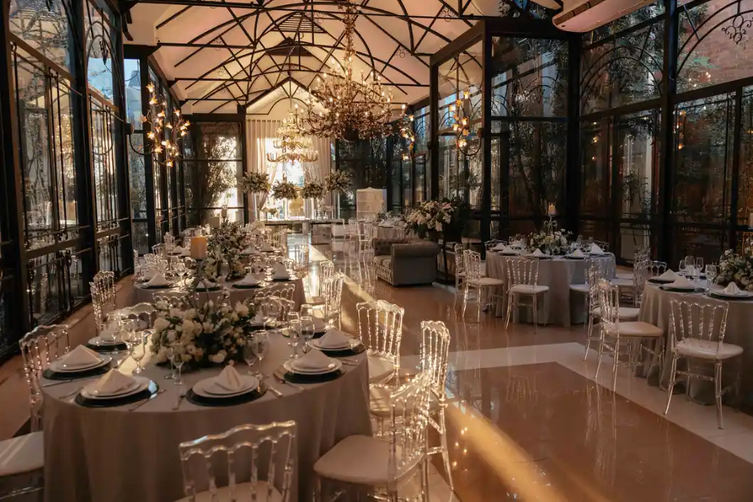 Salão de eventos decorado para casamento com mesas postas em estilo elegante, cadeiras com capas claras, arranjos florais e lustres iluminados sob uma estrutura de vidro.