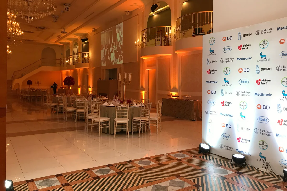 Salão de eventos decorado com iluminação amarela, mesas com toalhas verdes e arranjos florais vermelhos, cadeiras brancas e painel com logos de empresas patrocinadoras