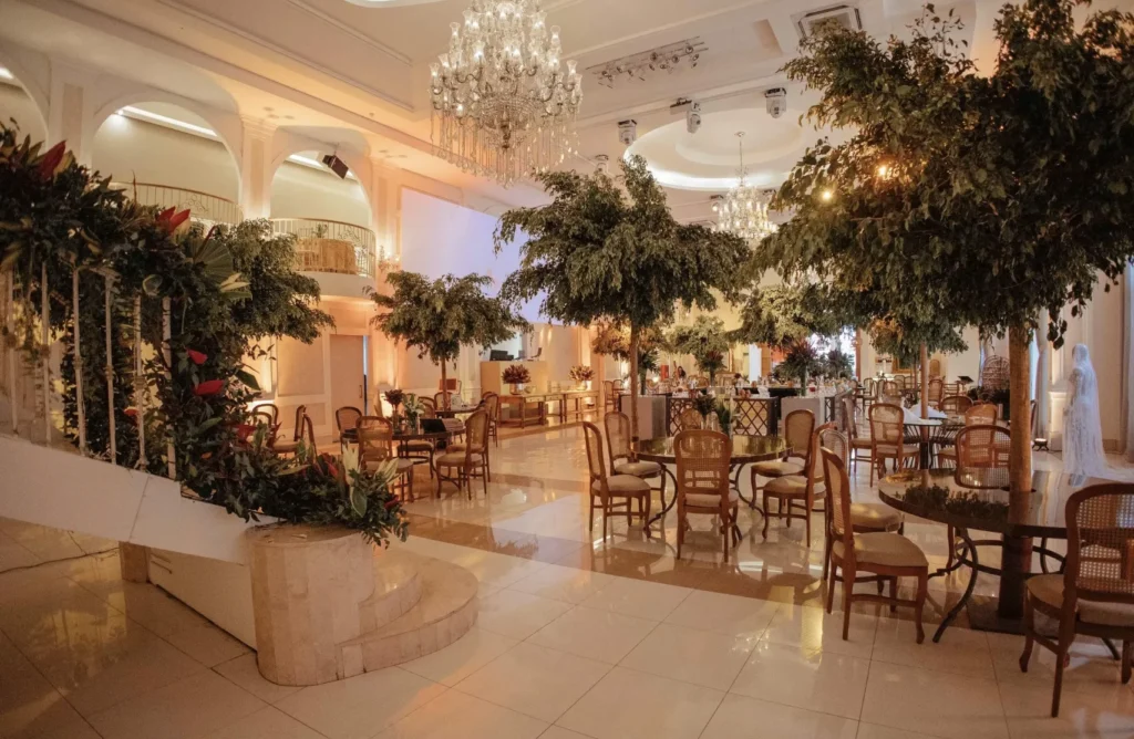 Salão de eventos elegante com lustres de cristal, árvores decorativas e mesas com cadeiras de madeira em ambiente iluminado